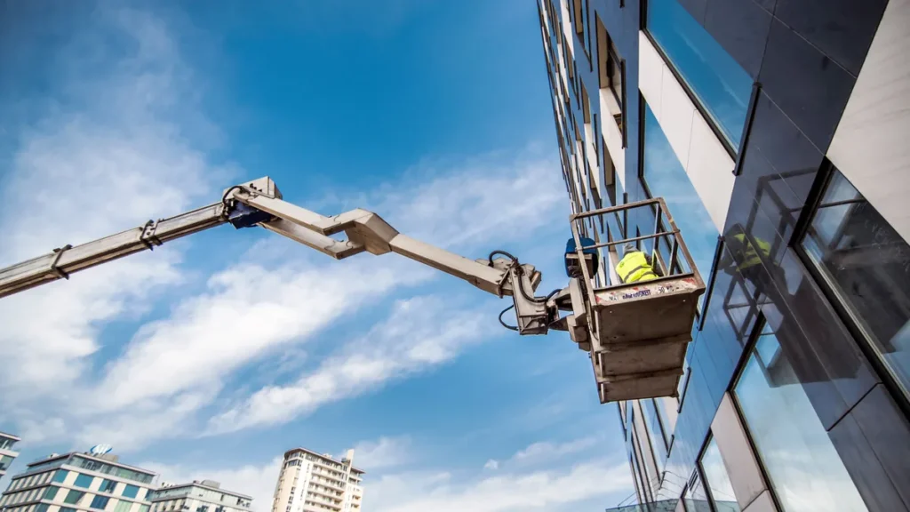 Trabalhador em plataforma elevatória articulada junto à fachada de um prédio, com céu azul ao fundo