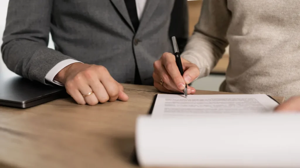O que posso comprar com o consórcio de imóveis? Pessoa assina um contrato com caneta sobre uma mesa de madeira, ao lado de outra pessoa de terno