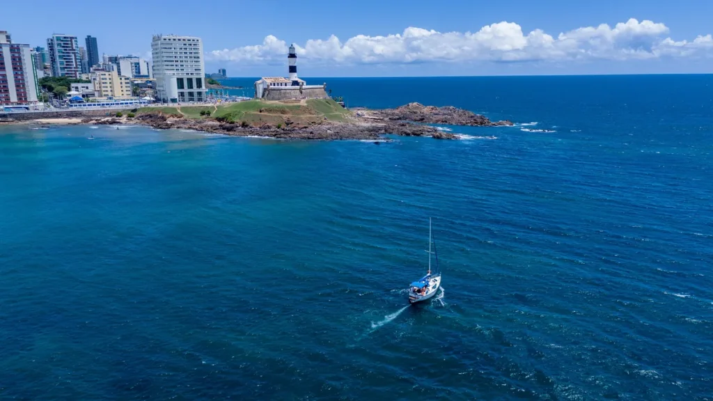 Planeje sua viagem de ônibus para Salvador! Vista aérea do Farol da Barra, em Salvador, com um veleiro navegando no mar azul