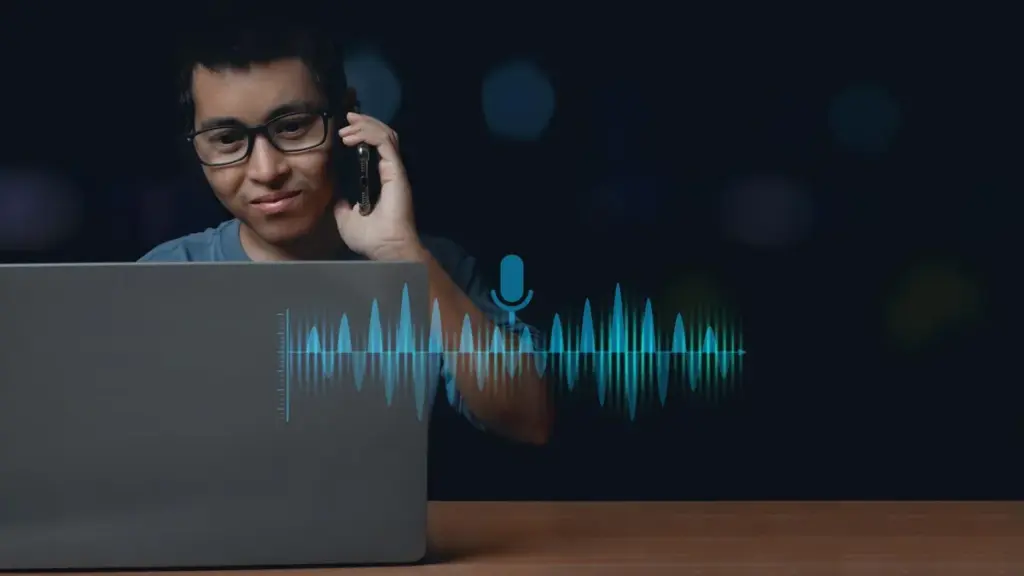 De Ouro Preto, healthtech amplia IA por voz para novos mercados Jovem de óculos fala ao celular diante do notebook, com uma onda sonora e um ícone de microfone em destaque