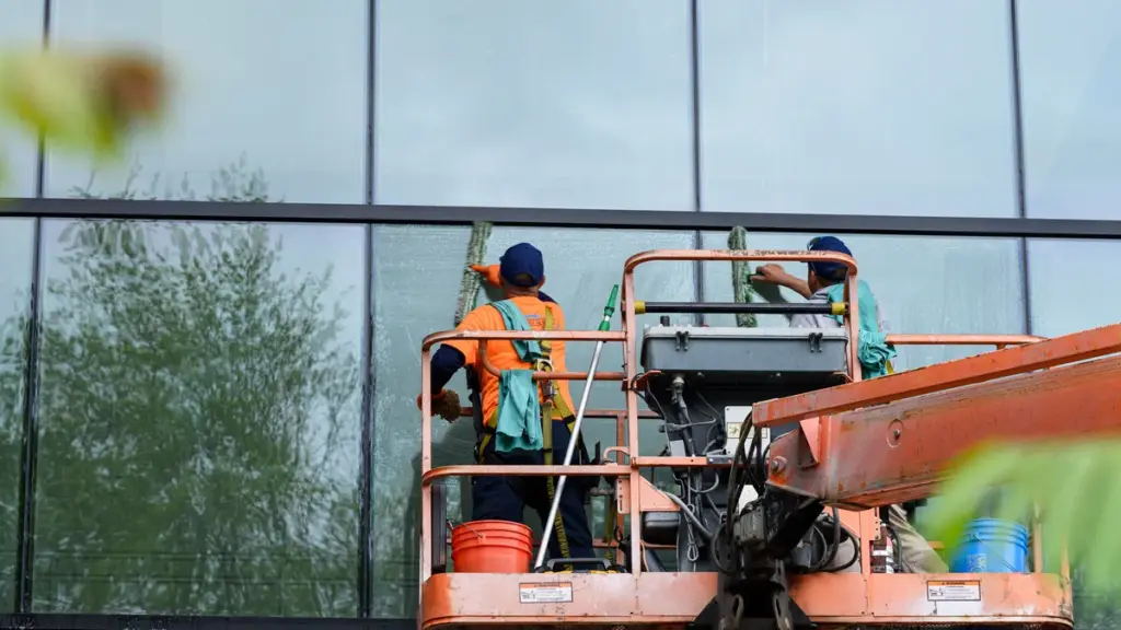 Dois trabalhadores limpam a fachada de vidro de um prédio em cima de uma plataforma elevatória laranja